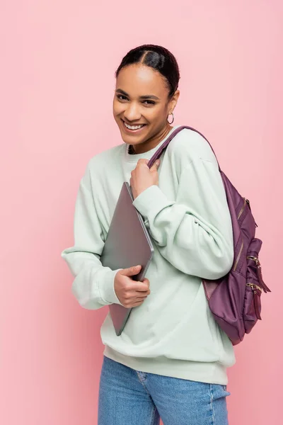 Estudiante afroamericano satisfecho con la mochila que sostiene el ordenador portátil aislado en rosa - foto de stock