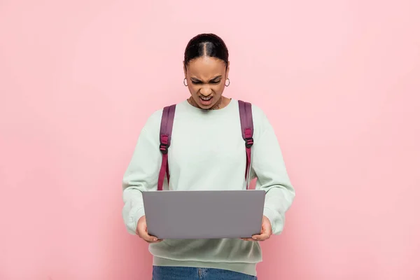Angry african american student with backpack using laptop isolated on pink — Stock Photo