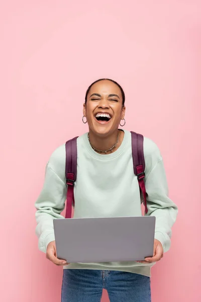 Feliz africano americano estudiante con mochila celebración portátil y riendo aislado en rosa - foto de stock
