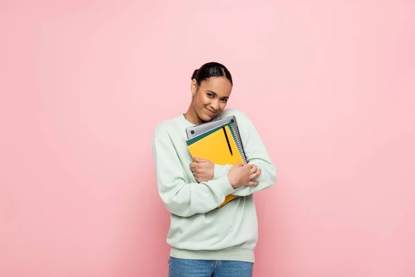 Estudiante afroamericano feliz en sudadera que sostiene el ordenador portátil y los cuadernos aislados en rosa - foto de stock