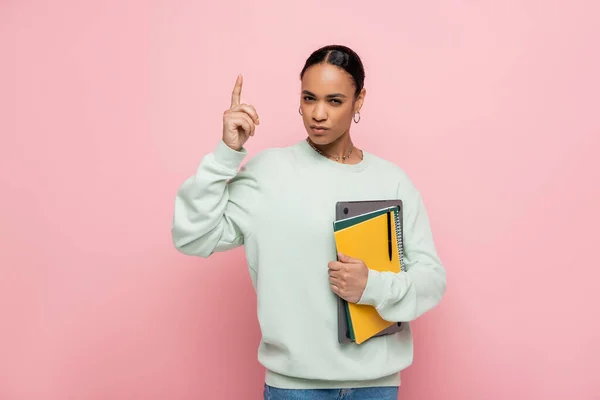 Estudiante afroamericano inteligente sosteniendo el ordenador portátil y los cuadernos mientras que tiene idea aislada en rosa - foto de stock