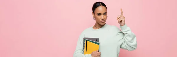 Estudiante afroamericano inteligente que sostiene el ordenador portátil y los suministros de estudio mientras que tiene idea aislada en rosa, bandera - foto de stock