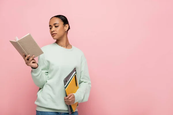 Розумний афроамериканський студент в сорочці читає книгу, тримаючи ноутбук і блокноти ізольовані на рожевому — стокове фото