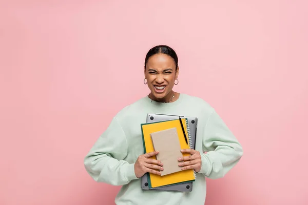 African american student in sweatshirt holding laptop and study supplies while grinning isolated on pink — Stock Photo