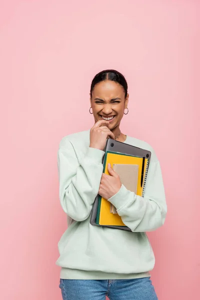 Cheerful african american student in sweatshirt holding laptop and study supplies isolated on pink — Stock Photo