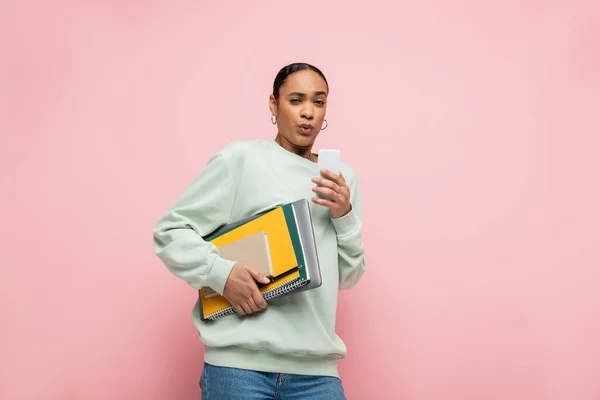 Estudante afro-americano tenso em suéter segurando suprimentos de estudo e smartphone isolado em rosa — Fotografia de Stock