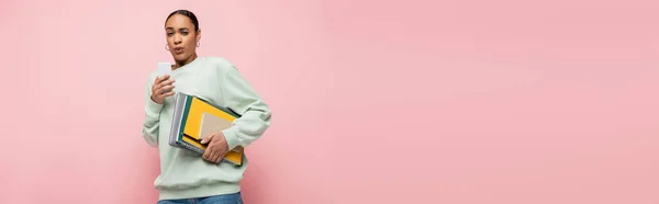Estudante afro-americano tenso em suéter segurando suprimentos de estudo e usando smartphone isolado em rosa, banner — Fotografia de Stock