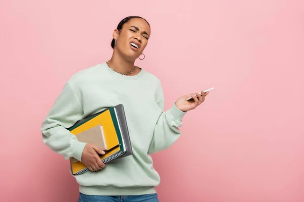Displeased african american student in sweatshirt holding gadgets and study supplies while whining isolated on pink — Stock Photo