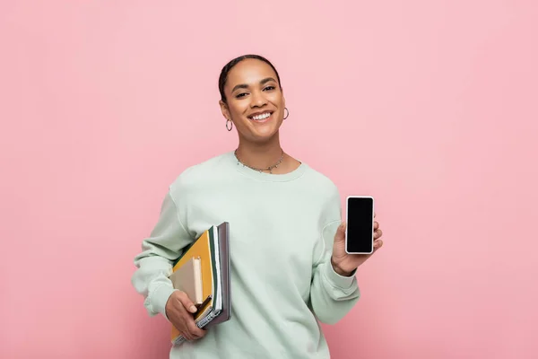 Estudiante afroamericano sonriente en sudadera con suministros de estudio y teléfono inteligente con pantalla en blanco aislado en rosa - foto de stock