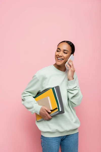 Joyeux étudiant afro-américain en sweat-shirt tenant des fournitures d'étude et parlant sur smartphone isolé sur rose — Photo de stock