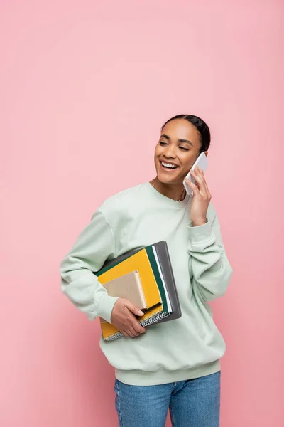 Cheerful african american student in sweatshirt holding study supplies and talking on smartphone isolated on pink — Stock Photo