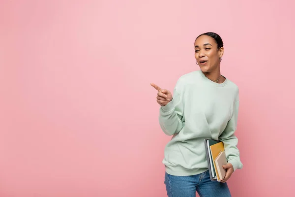 African american student in sweatshirt holding study supplies and making fun while pointing with finger isolated on pink — Stock Photo