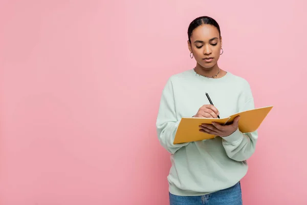 Гарненька афроамериканська студентка в сорочці робить нотатки під час вивчення ізольовано на рожевому — стокове фото