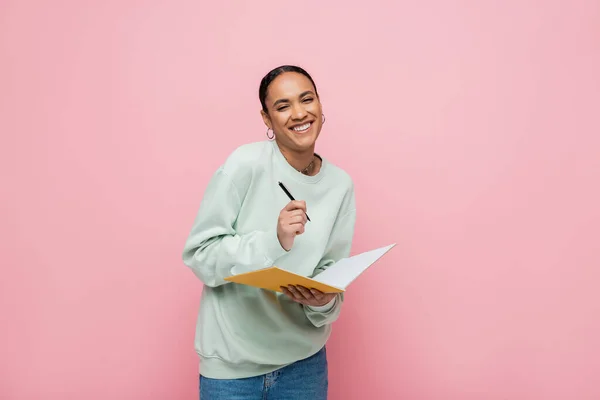 Alegre estudante afro-americano em suéter segurando caneta e notebook isolado em rosa — Fotografia de Stock