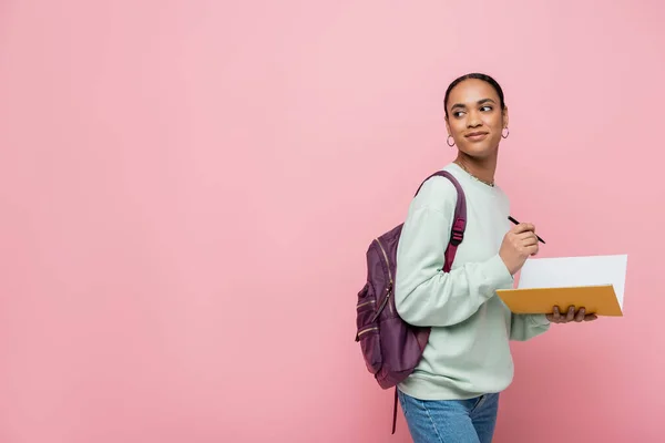 Estudiante afroamericano sonriente con mochila que sostiene la pluma y el cuaderno aislado en rosa - foto de stock