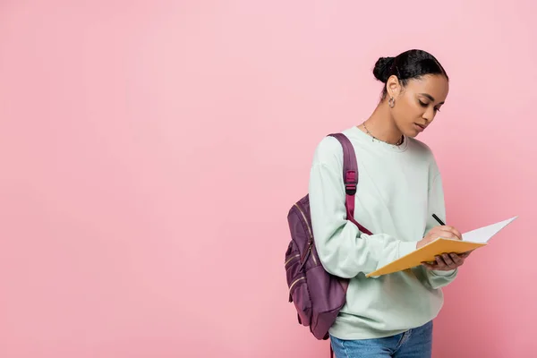 Brunette african american student taking notes while holding notebook and pen isolated on pink — Stock Photo