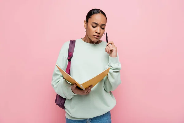 Frustrierte afrikanisch-amerikanische Studentin hält Notizbuch und Stift in der Hand, während sie mit Rucksack isoliert auf rosa steht — Stockfoto