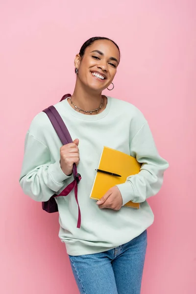Веселий афроамериканський студент тримає блокнот і ручку, стоячи з рюкзаком ізольовано на рожевому — стокове фото