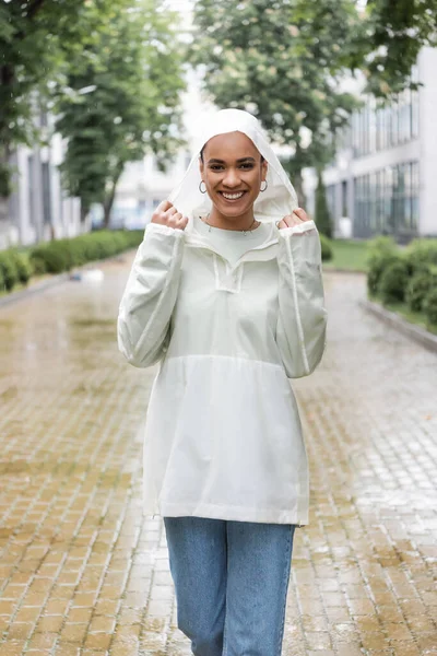 Cheerful african american woman in waterproof raincoat standing under rain outdoors — Stock Photo