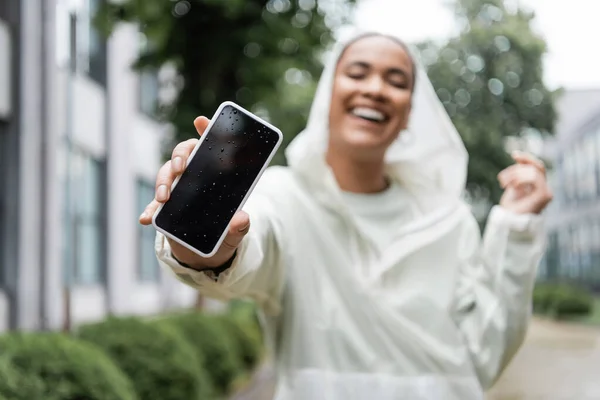 Alegre mujer afroamericana en impermeable con capucha sosteniendo teléfono inteligente húmedo con pantalla en blanco - foto de stock