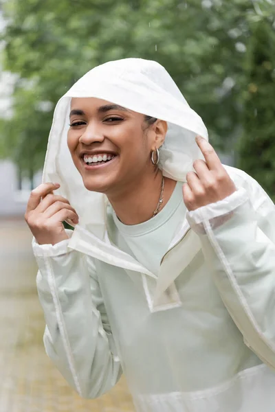 Femme afro-américaine ravie en imperméable imperméable capuche ajustable pendant la pluie — Photo de stock