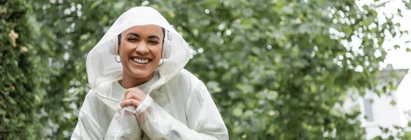 Lächelnde Afroamerikanerin in wasserdichtem Regenmantel und kabellosen Kopfhörern, die unter Regen steht, Banner — Stockfoto