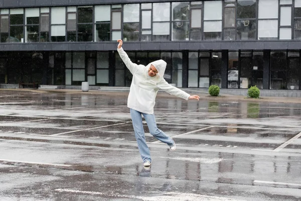 Comprimento total de mulher americana africana feliz em capa impermeável e jeans se divertindo durante a chuva — Fotografia de Stock