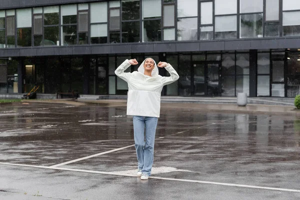 Comprimento total de mulher afro-americana alegre em impermeável capa de chuva e jeans se divertindo durante a chuva — Fotografia de Stock