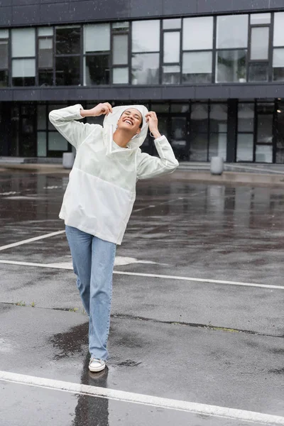 Повна довжина задоволеної афроамериканської жінки в водонепроникному плащі і джинсах, розважаючись під час дощу — стокове фото