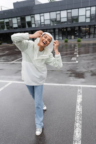 Longitud completa de la mujer afroamericana emocionada en impermeable impermeable y jeans divertirse durante la lluvia - foto de stock