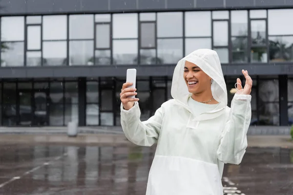 Happy african american woman in waterproof raincoat taking selfie on smartphone during rain — Stock Photo