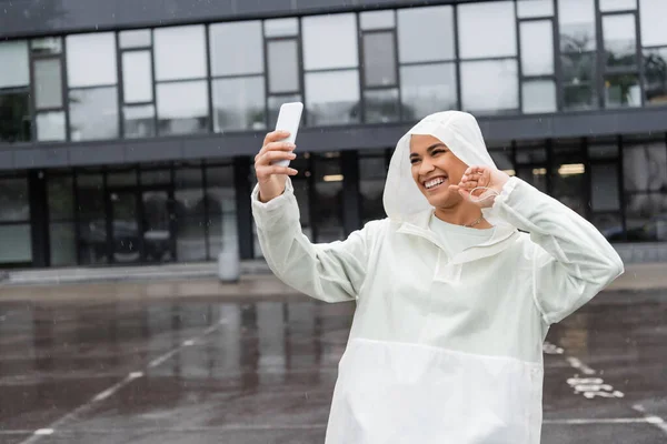 Американка в водонепроницаемом плаще делает селфи на смартфоне во время дождя — стоковое фото