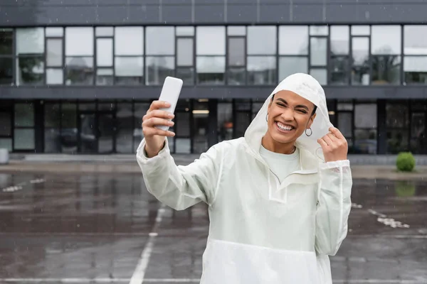 Cheerful african american woman in waterproof raincoat taking selfie on smartphone during rain — Stock Photo