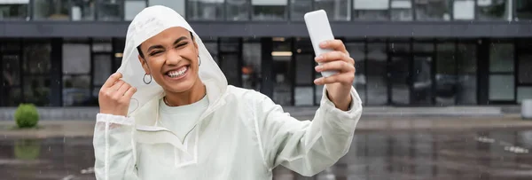 Alegre mujer afroamericana en impermeable tomando selfie en teléfono inteligente durante la lluvia, bandera - foto de stock