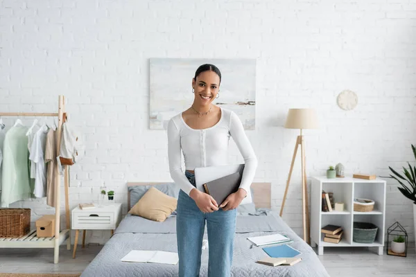 Cheerful african american student standing with laptop and book in modern bedroom — Stock Photo