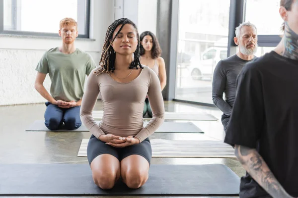 Mujer afroamericana joven meditando en Thunderbolt posan cerca de grupo en estudio de yoga - foto de stock