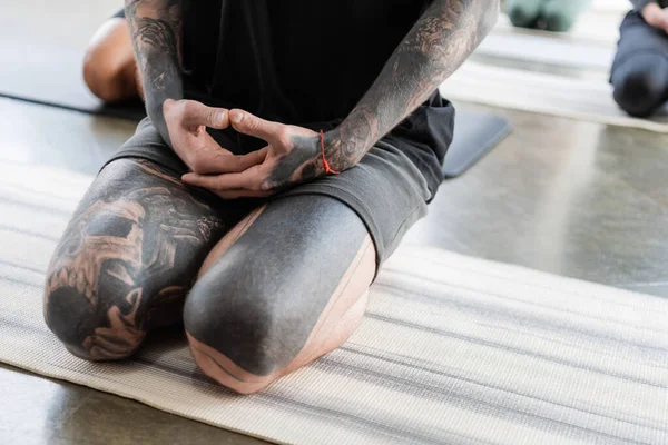 Обрезанный вид на татуированного человека, сидящего в Thunderbolt asana на коврике для йоги в студии — стоковое фото