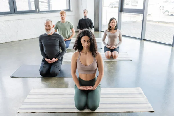 Personas multiétnicas meditando con los ojos cerrados en Thunderbolt asana en estudio de yoga - foto de stock