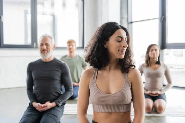 Sonriente mujer de Oriente Medio meditando cerca de personas borrosas en clase de yoga - foto de stock