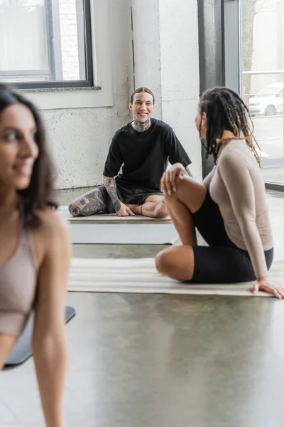 Lächelnde gemischtrassige Menschen reden, während sie im Yoga-Kurs auf Matten sitzen — Stockfoto