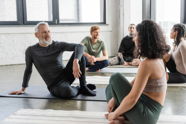 Hombre maduro positivo hablando con la mujer de Oriente Medio en clase de yoga - foto de stock