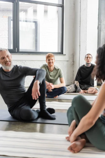 Cheerful men talking to blurred woman on mat in yoga class — Stock Photo