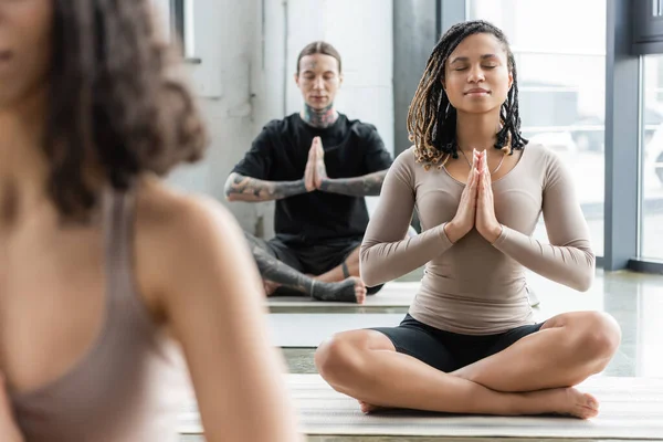 Femme afro-américaine méditant avec anjali mudra près du groupe flou dans le cours de yoga — Photo de stock