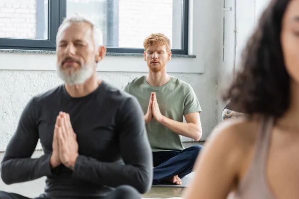 Pelirroja meditando con anjali mudra cerca de personas borrosas en clase de yoga - foto de stock