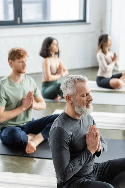 Hombre maduro sentado en anjali mudra cerca de grupo multiétnico borroso en clase de yoga - foto de stock
