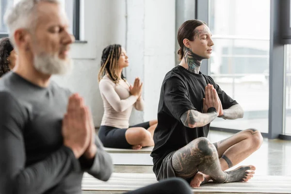 Татуйований чоловік практикує анжалі мудра поблизу багатоетнічних людей в групі йоги — стокове фото