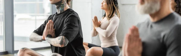 Hombre tatuado haciendo anjali mudra cerca de grupo interracial en clase de yoga, pancarta - foto de stock
