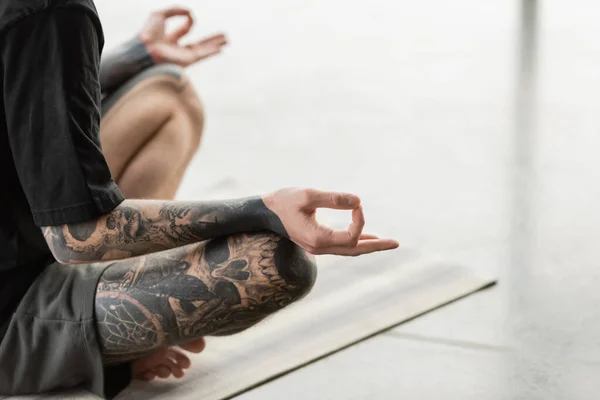 Vista ritagliata del giovane uomo tatuato che pratica mudra gyan sul tappeto in studio di yoga — Foto stock