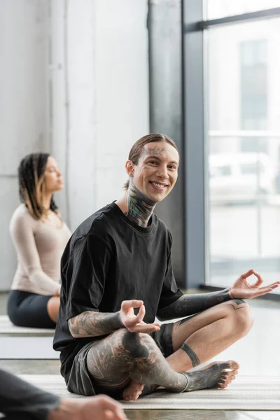 Позитивный татуированный мужчина, смотрящий в камеру во время занятий гианской мудрой в студии йоги — стоковое фото