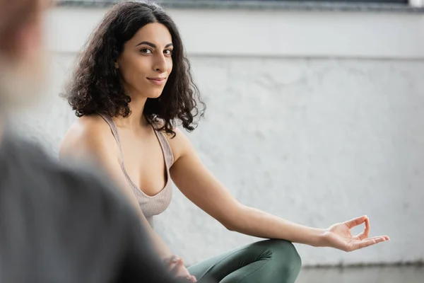 Sonriente mujer de Oriente Medio en ropa deportiva haciendo gyan mudra y mirando a la cámara en clase de yoga - foto de stock